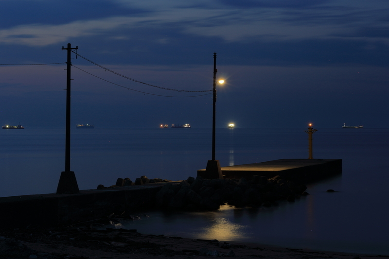 四日市市の楠漁港にある南側の突堤と朝の海の様子を撮影した写真①突堤には電灯と赤い小さな灯台があり、沖合を多くの大型船が航行しています。海は静か。